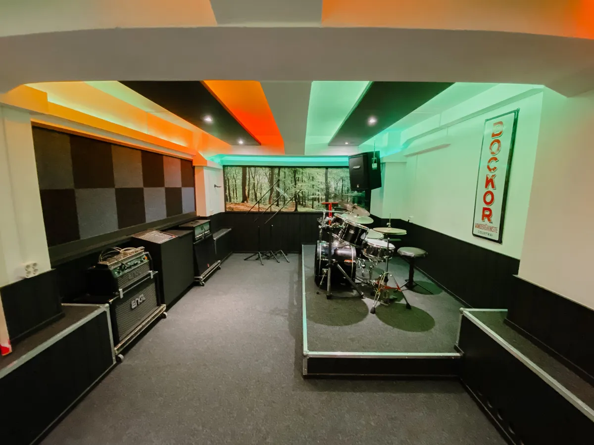 Eigen Jam-oefenstudio met drumstel op verhoging, gitaar- en basversterkers, microfoonstatieven en vrije speelvloer
