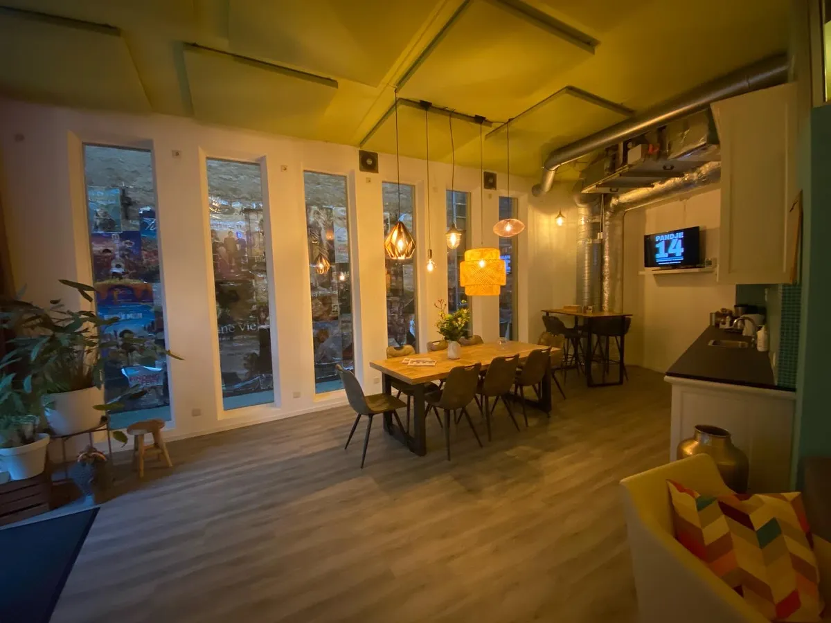 Huiskamer van PANDJE14 met hoge ramen, houten tafel, zitplekken, keuken en hanglampen